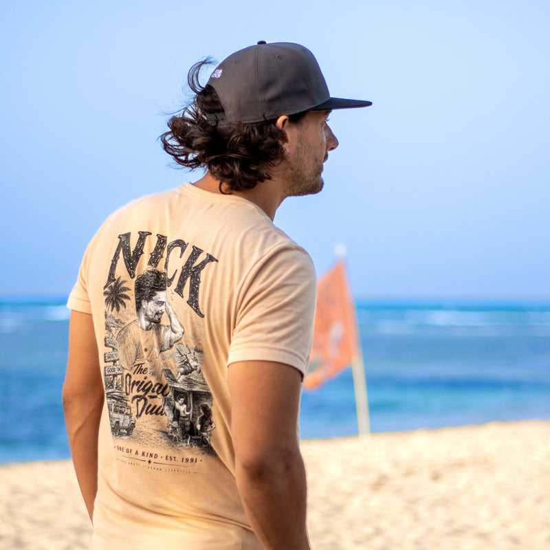 A man standing on a sandy beach wearing a custom Gone Salty graphic t-shirt with a custom print on the back, a dark gray Gone Salty baseball cap, looking out towards the ocean.
