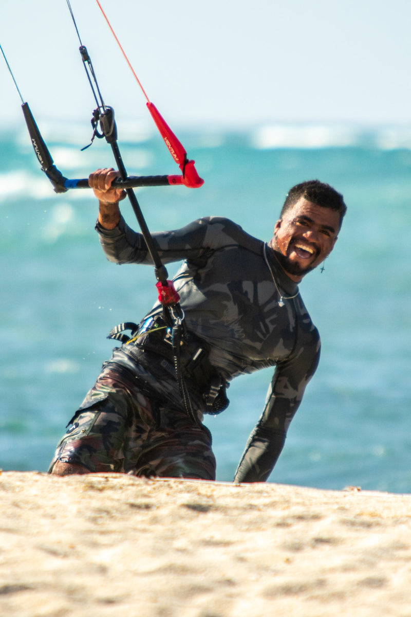 A man smiling and posing on a sandy beach while wearing a dark gray long-sleeve rash guard and camouflage pants, holding a kitesurfing bar with red and black straps against a backdrop of the ocean.