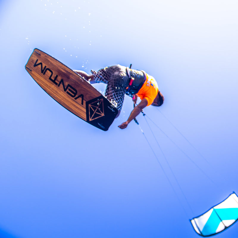 A kitesurfer in mid-air performing a trick against a clear blue sky, with a VENTUM board visible and part of a teal and white kite below.
