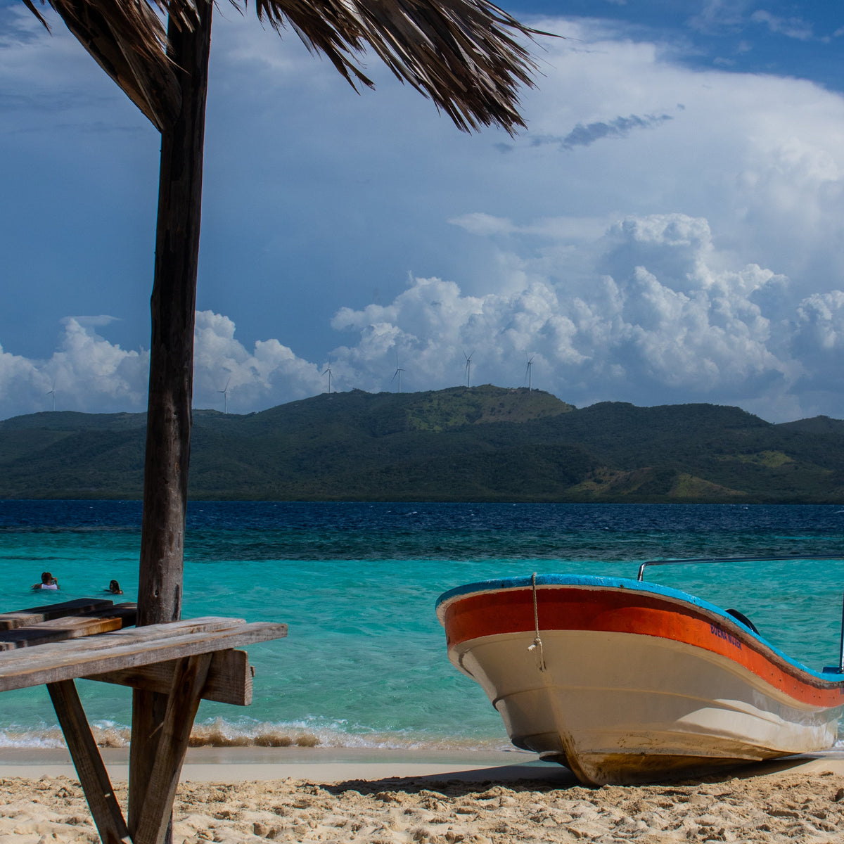 A tropical beach scene with a thatched umbrella in the foreground, a small white and orange boat resting on the sandy shore, turquoise water, distant green hills, and a blue sky with large white clouds.
